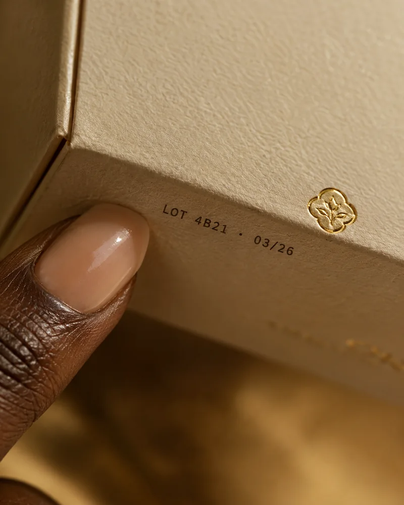 A fingertip beside the batch code and gold hallmark on a perfume box